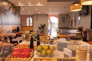 Un bufé con diversos alimentos, entre ellos frutas y postres, y un comedor con mesas y sillas.