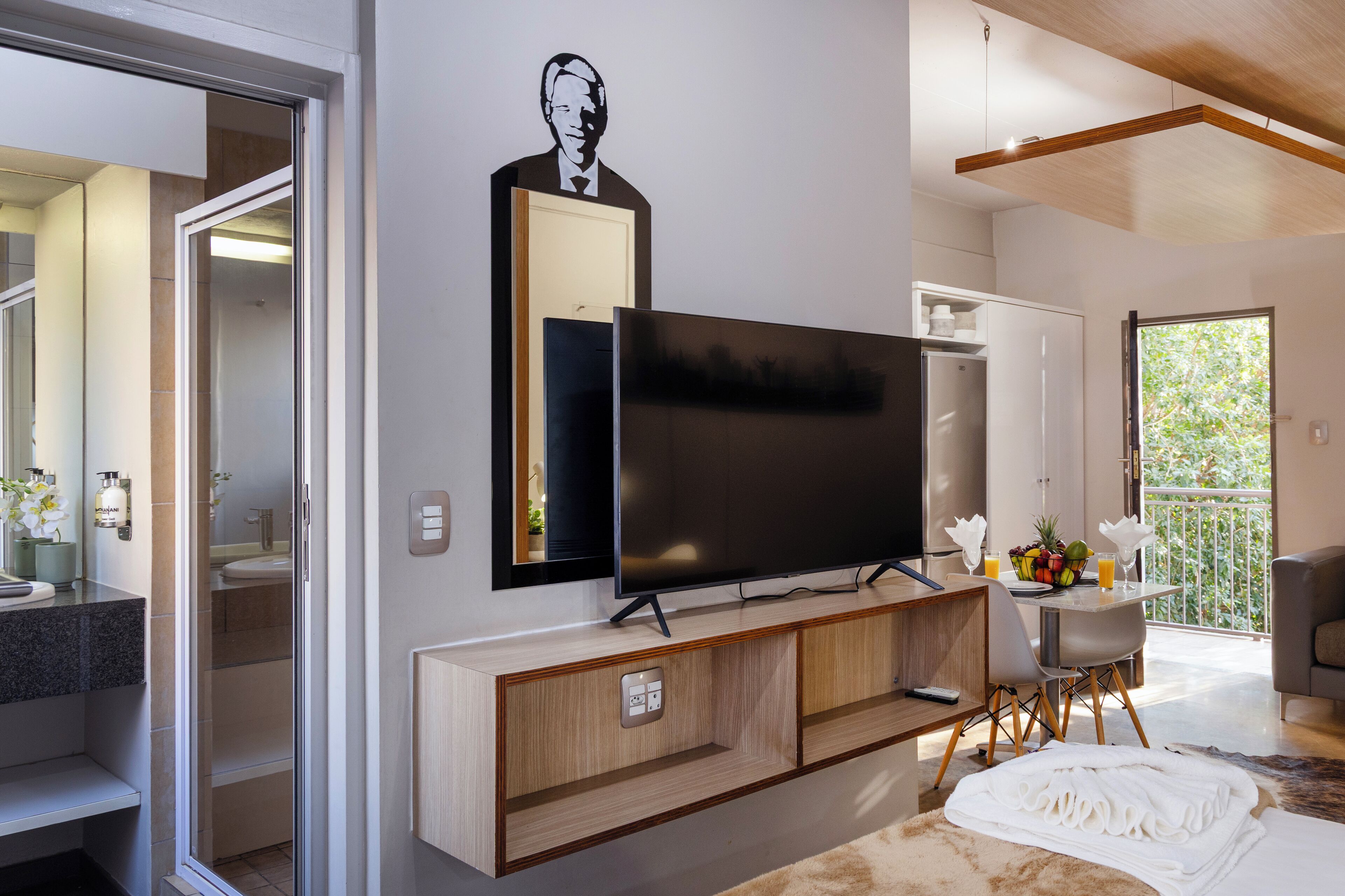 A modern living room with a flat-screen TV, a wooden entertainment unit, a dining area with a table and chairs, and a balcony with a view.