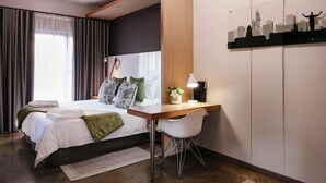 Ett modernt hotellrum med en säng, ett skrivbord, en stol och ett konstverk med en stadsbild på väggen.