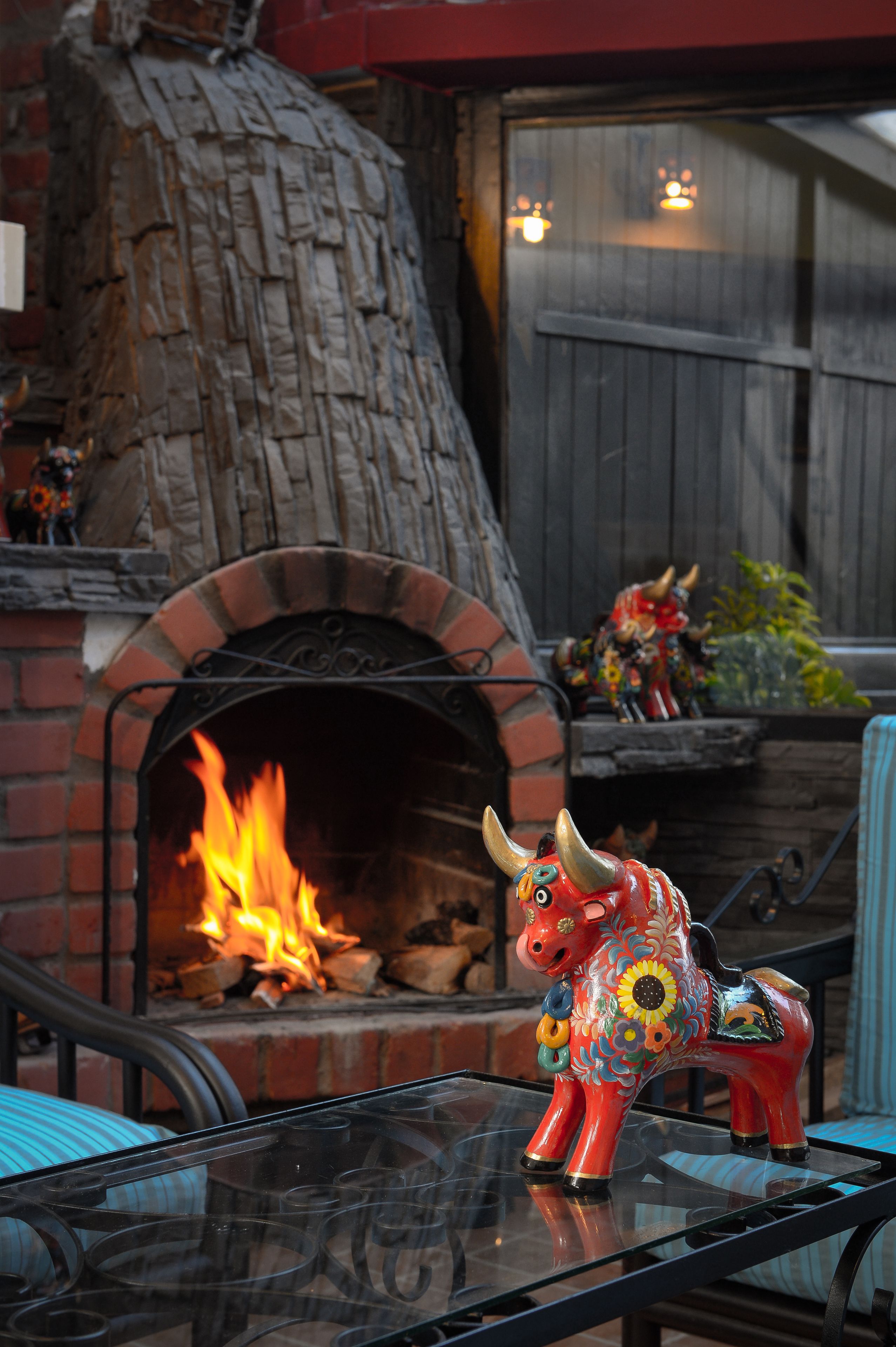 A rustic outdoor seating area with a brick fireplace, a decorative bull sculpture, and a glass table.