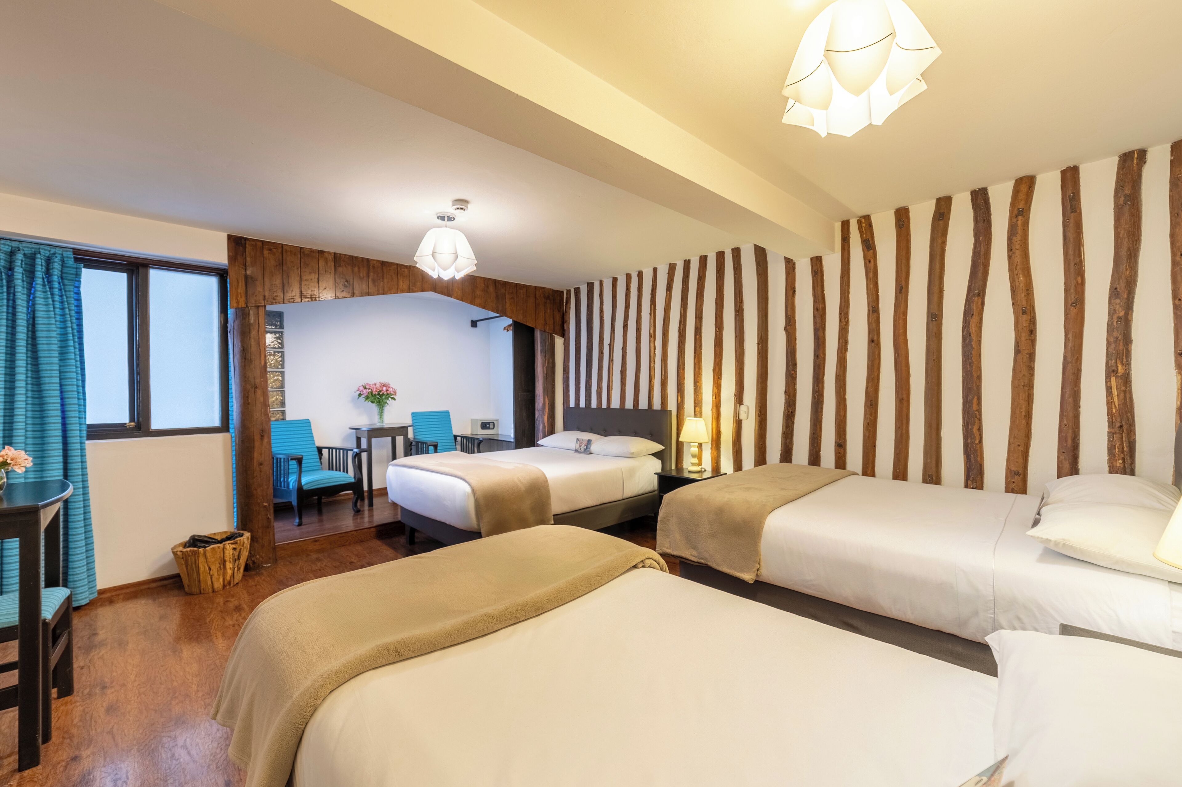 Ein Hotelzimmer mit zwei Betten, einem hölzernen Kopfteil, einem kleinen Schreibtisch mit Stuhl, einem Tisch mit Blumenvase und Blick ins Freie durch ein Fenster.
