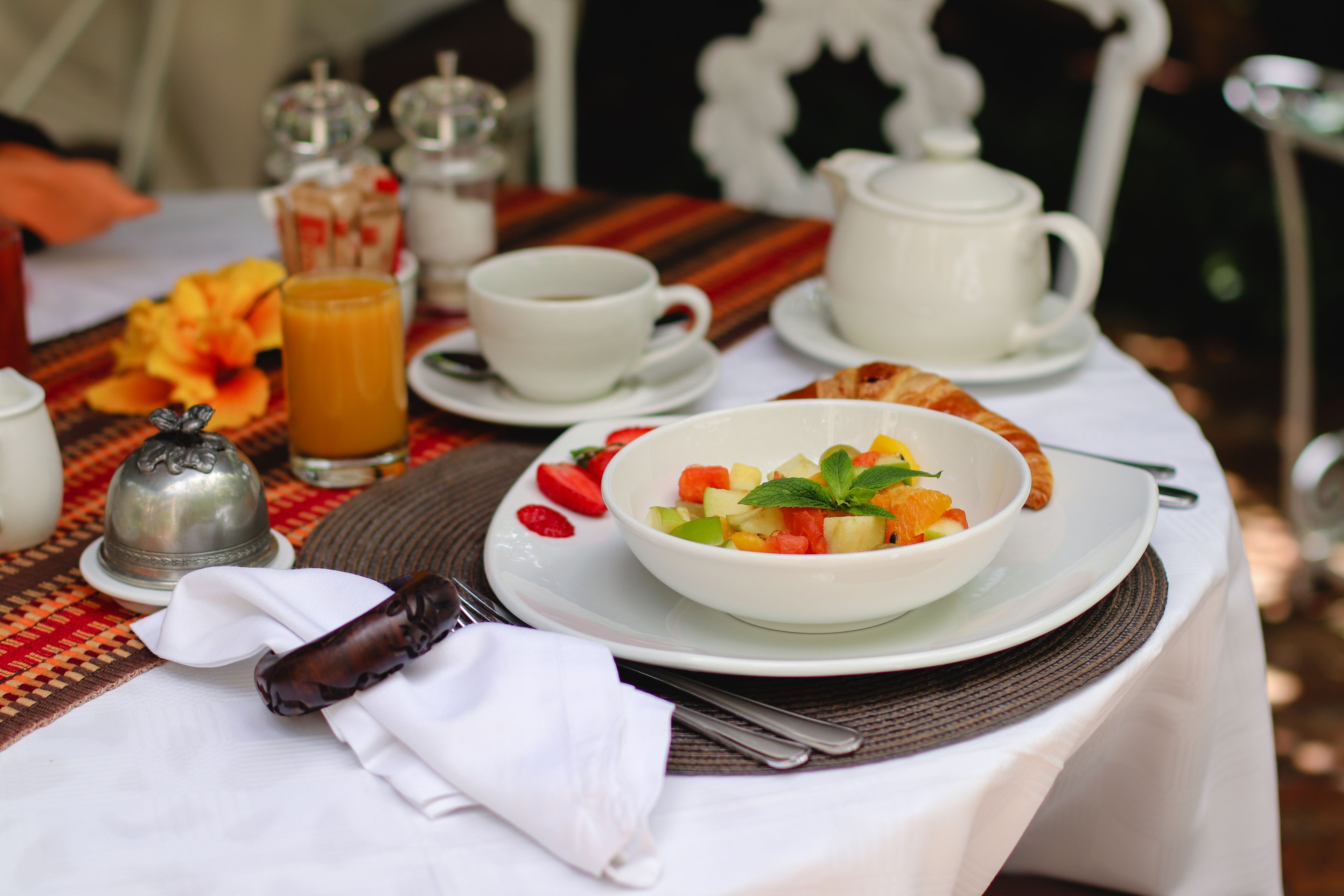 Un desayuno con una ensalada de frutas, una taza de café y un calentador de agua sobre una mesa con mantel a rayas.