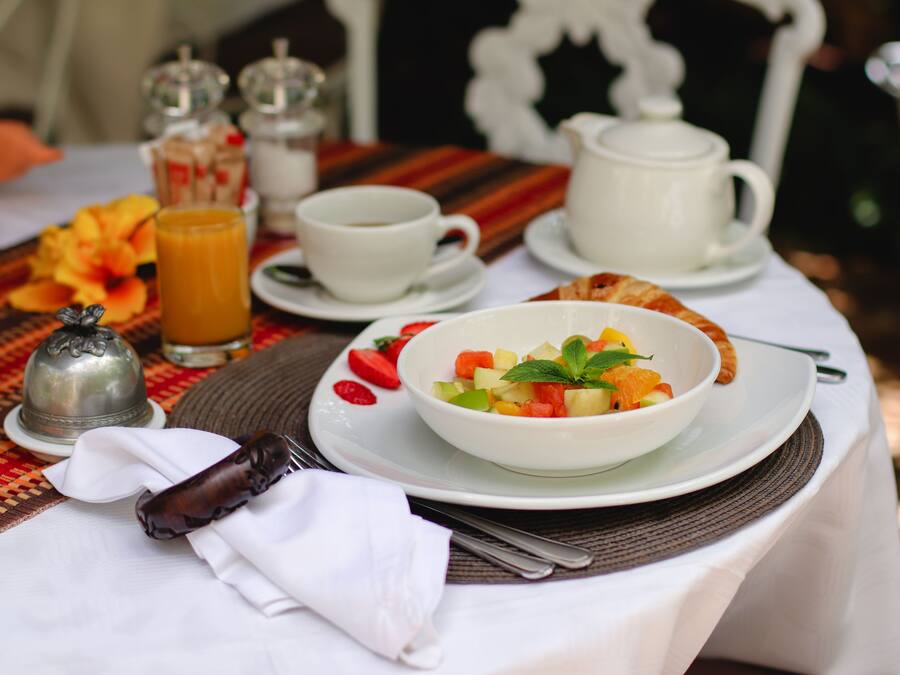 Un desayuno con una ensalada de frutas, una taza de café y un calentador de agua sobre una mesa con mantel a rayas.