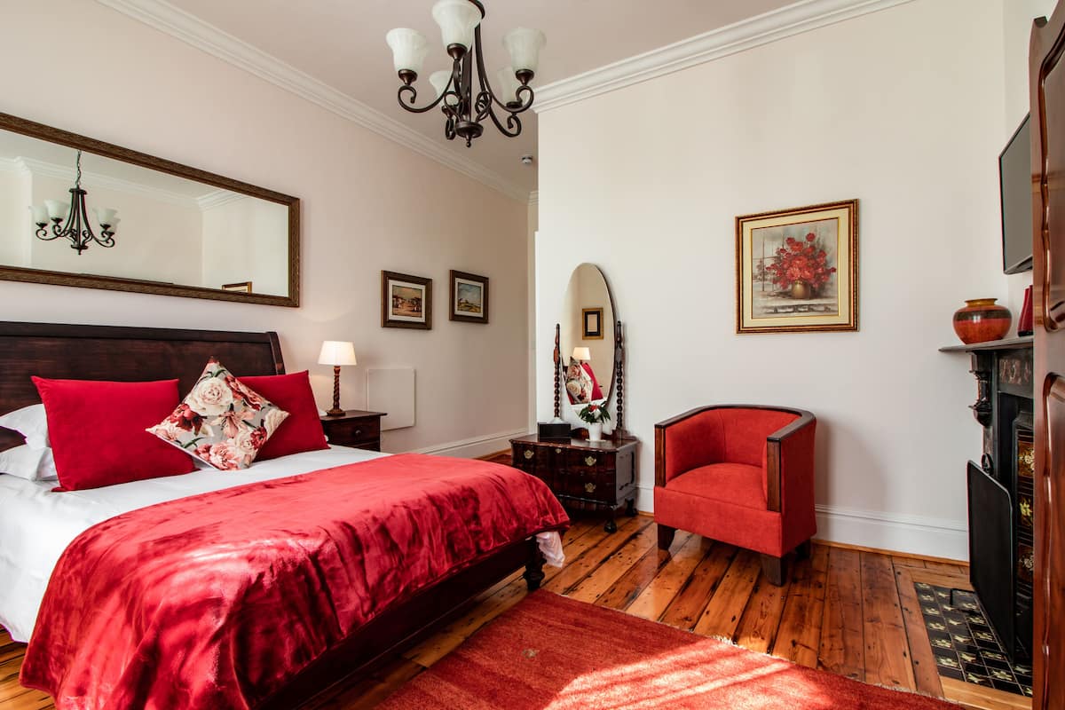 Ein Schlafzimmer mit einem großen Bett, einem roten Sessel, Holzboden, einem Spiegel und einem Kamin.