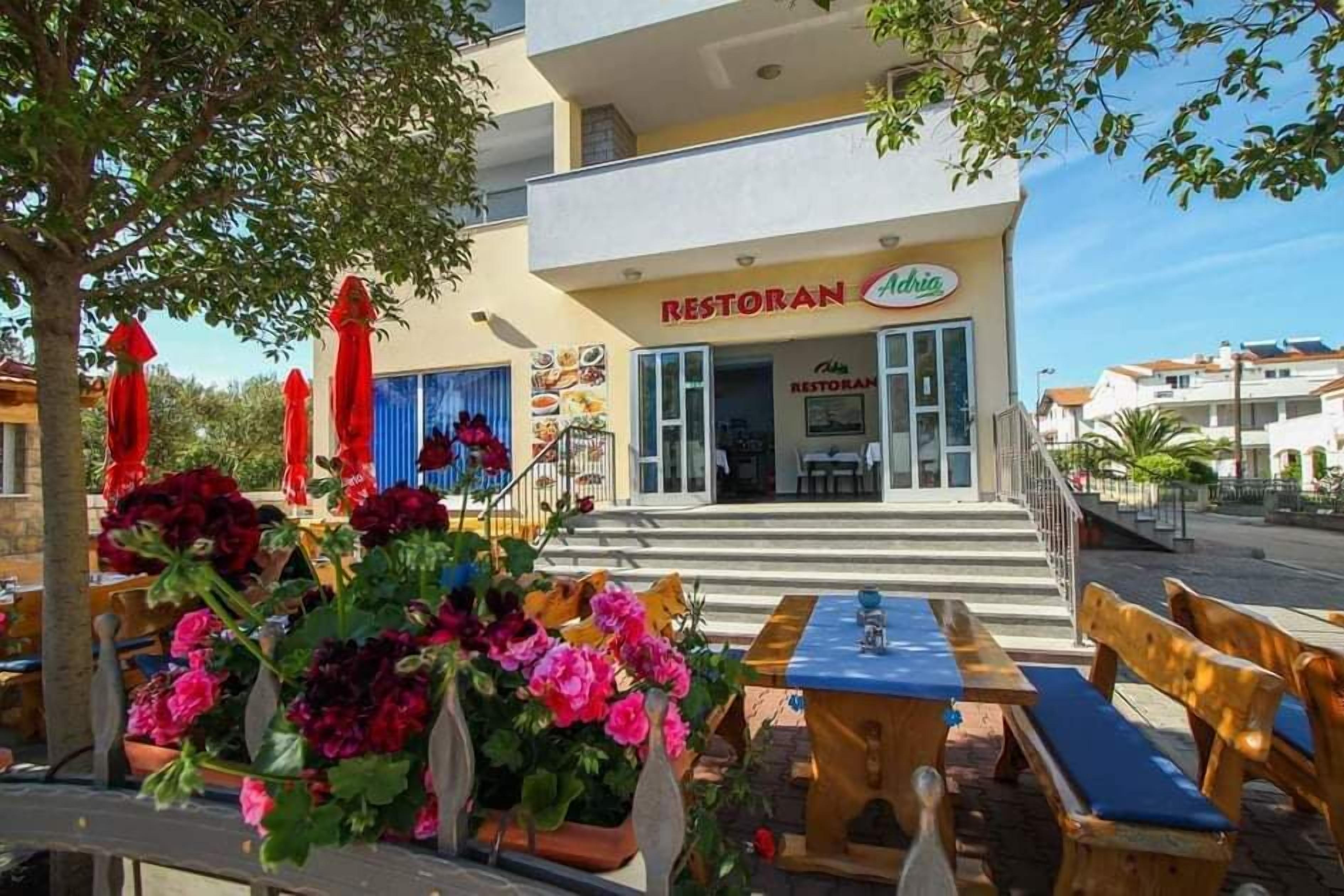 Egy szabadtéri ülőhely, ahol asztalok és padok találhatók, egy épület, amelyen „Restoran” felirat látható, és egy lépcső, amely a bejárathoz vezet.