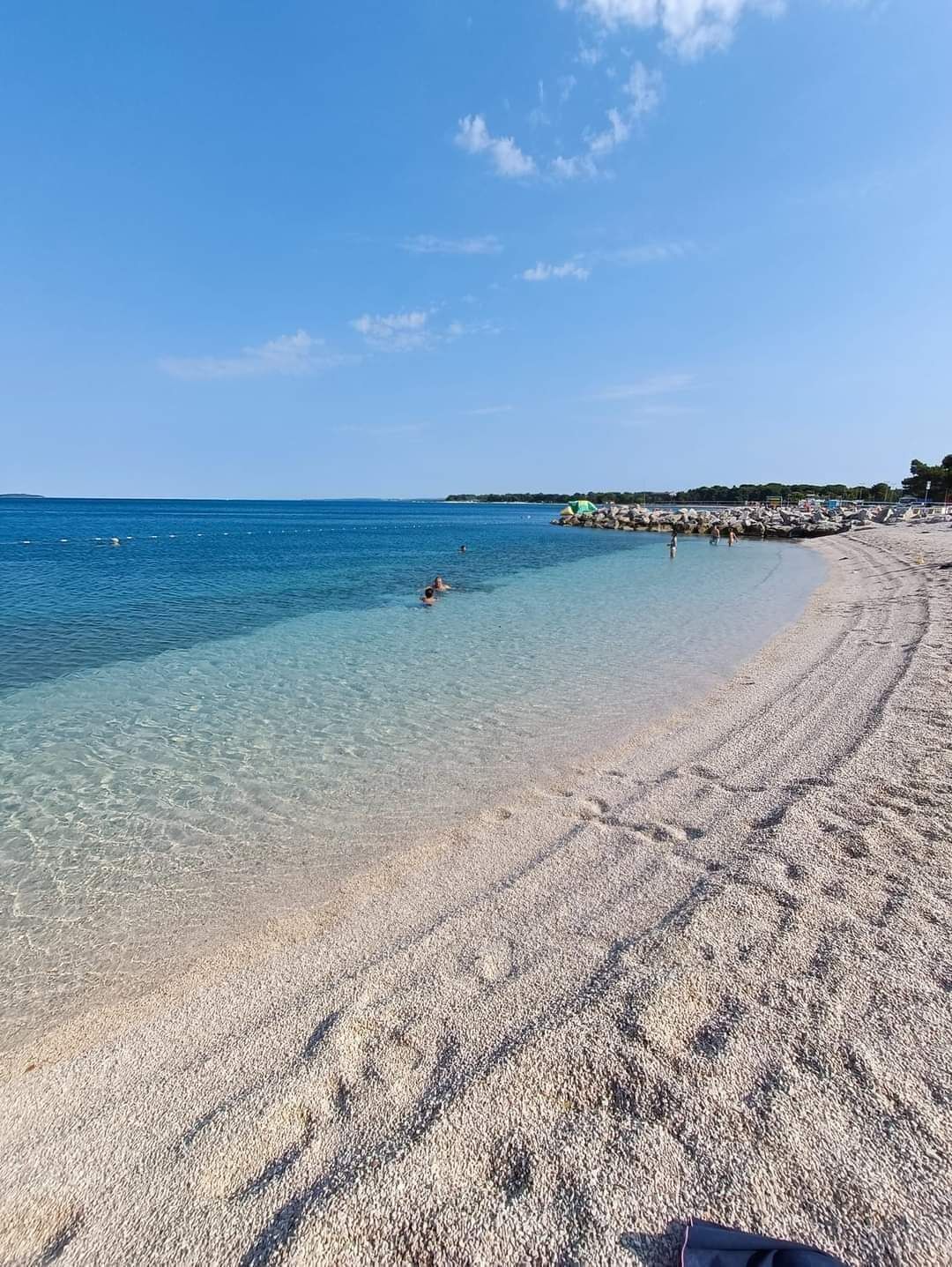 A beach with clear shallow waters, a pebbly shore, and a distant rocky outcrop.