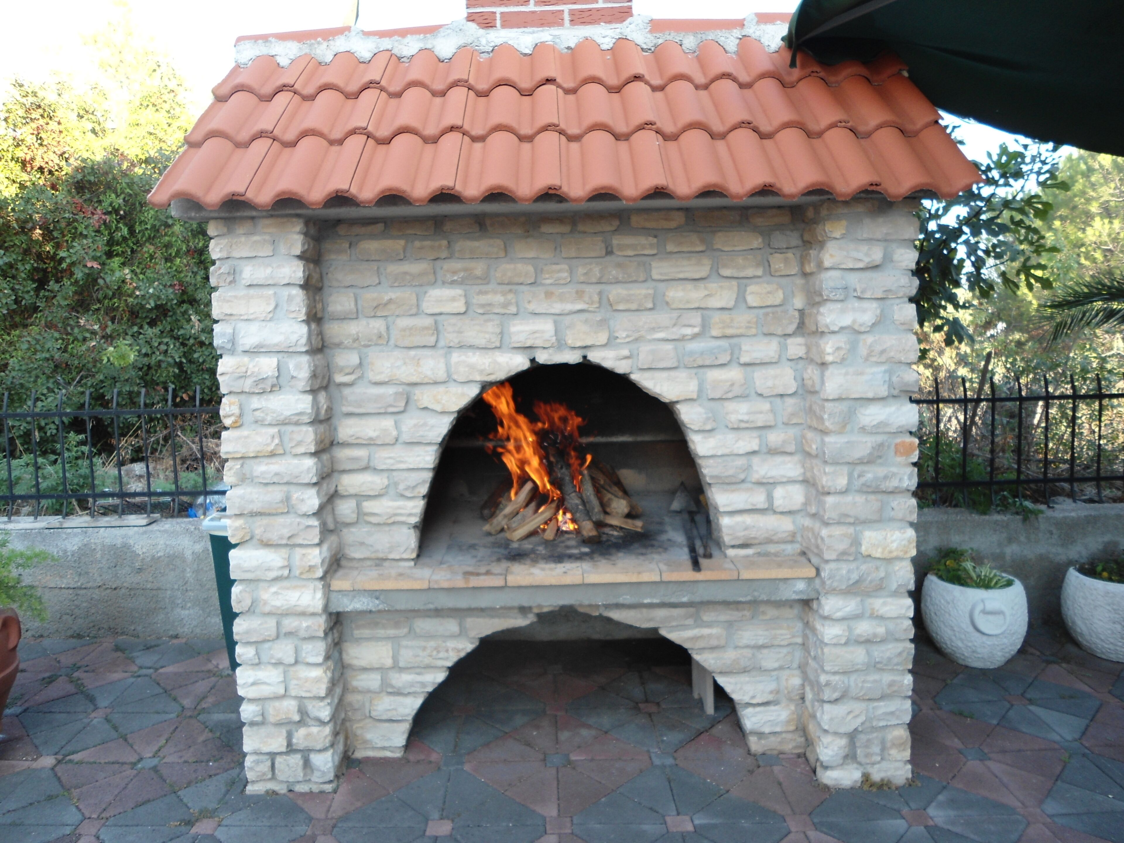 A stone-built outdoor pizza oven with a tiled roof, surrounded by a paved patio and a metal fence.