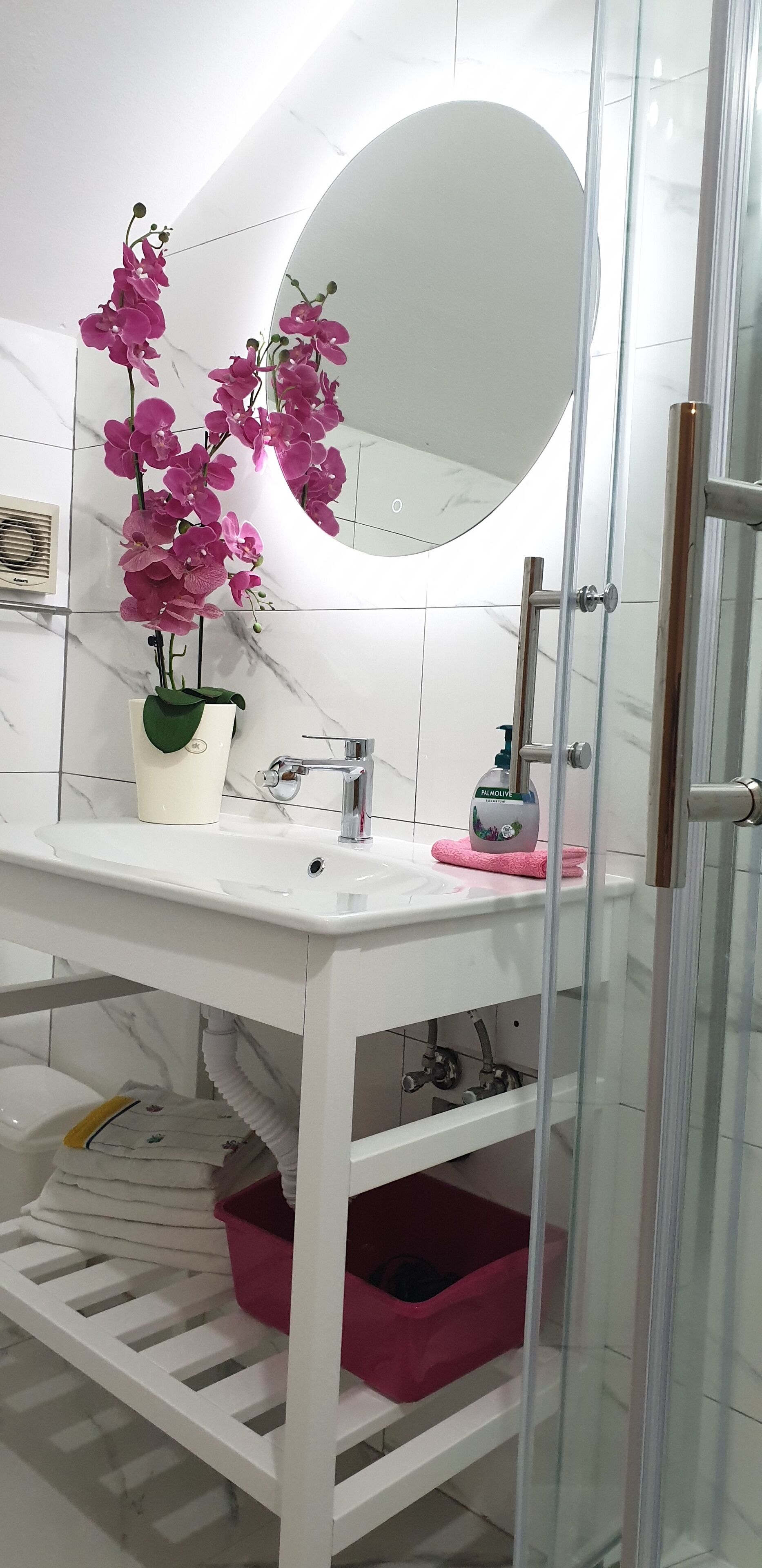 Une salle de bain avec un lavabo blanc, un grand miroir, une orchidée rose et une cabine de douche en verre.