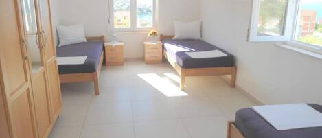 A room with two beds, a wooden wardrobe, a small table with a lamp, and a window with a view of buildings.