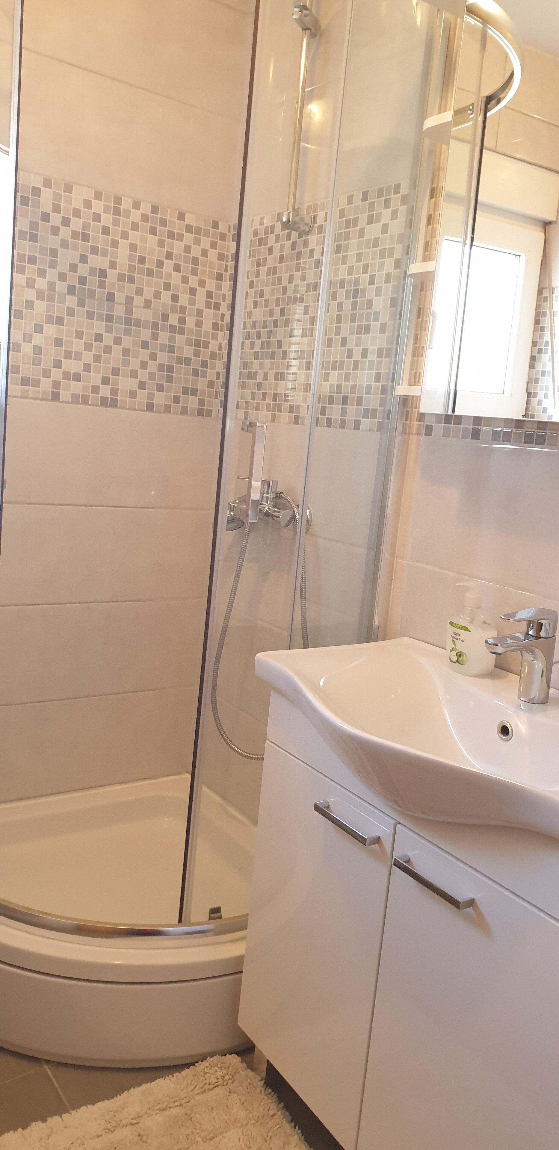 Ein modernes Badezimmer mit einer gläsernen Duschkabine, einem weißen Waschbecken mit Seifenspender und einer gefliesten Rückwand.