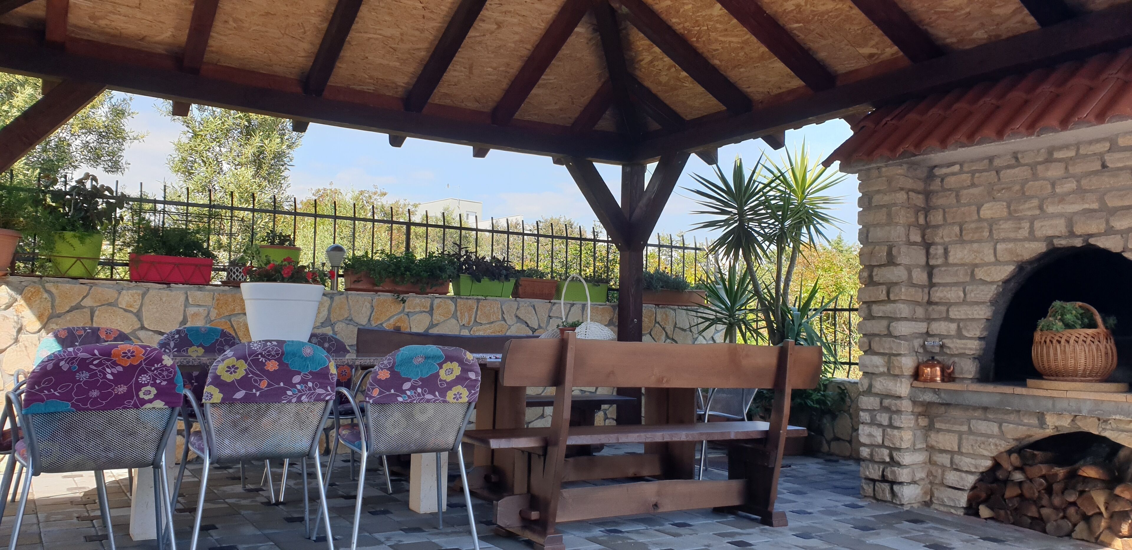 Eine Terrasse im Freien mit einer Steinsäule, einer Holzbank, Metallstühlen mit floralen Kissen und einem Kamin aus Ziegelsteinen.