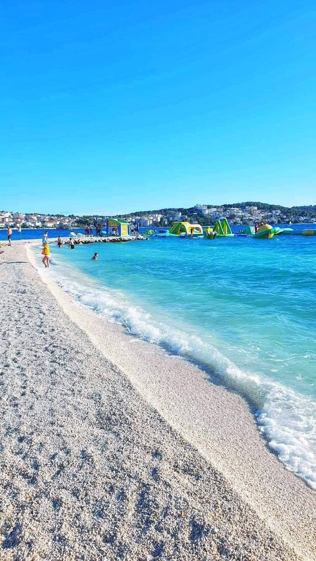 A beach with clear blue water, pebbly shore, and green water slides.