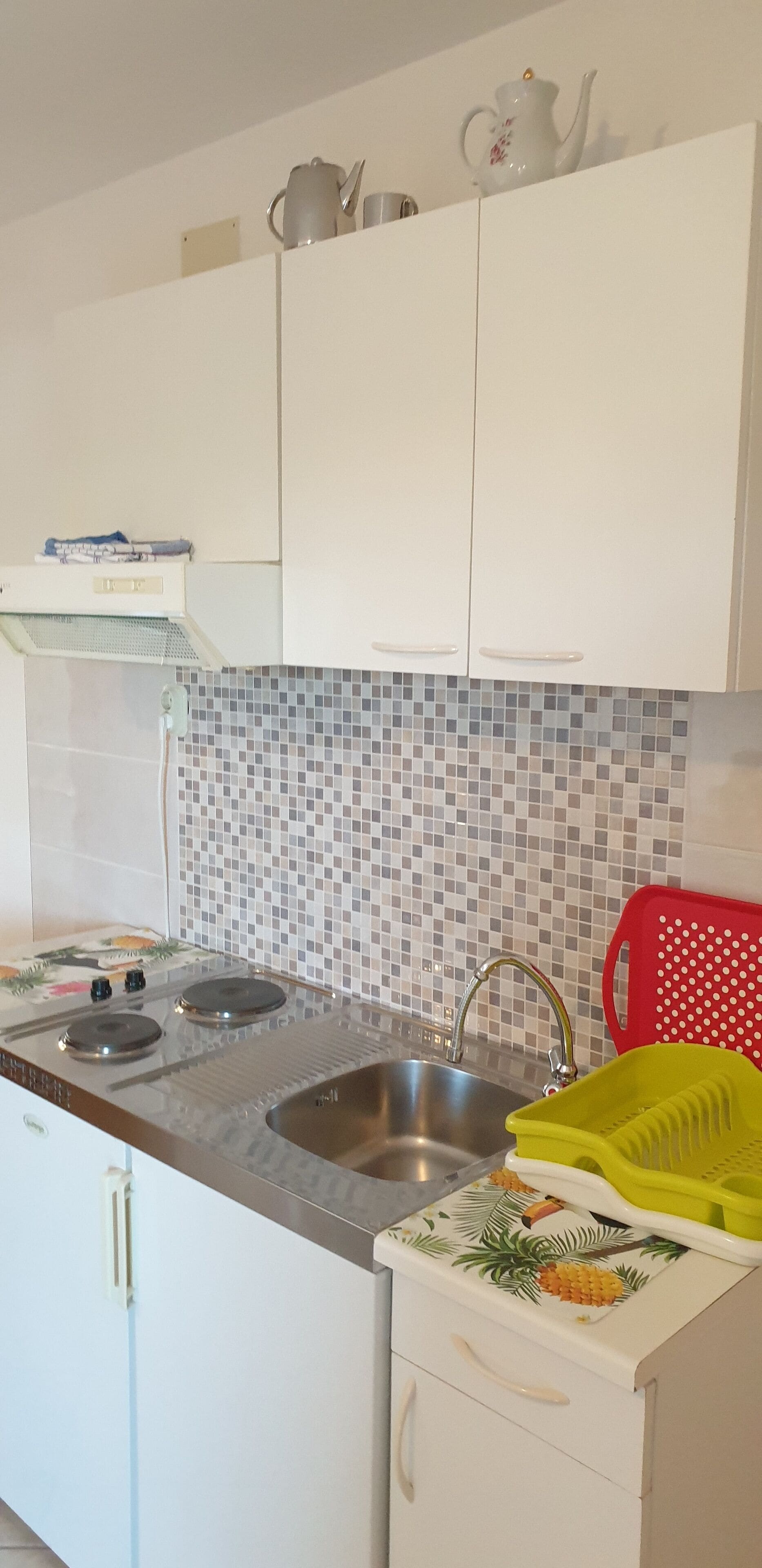 A kitchen with white cabinets, a stainless steel sink, a tiled backsplash, and a red plastic basket.