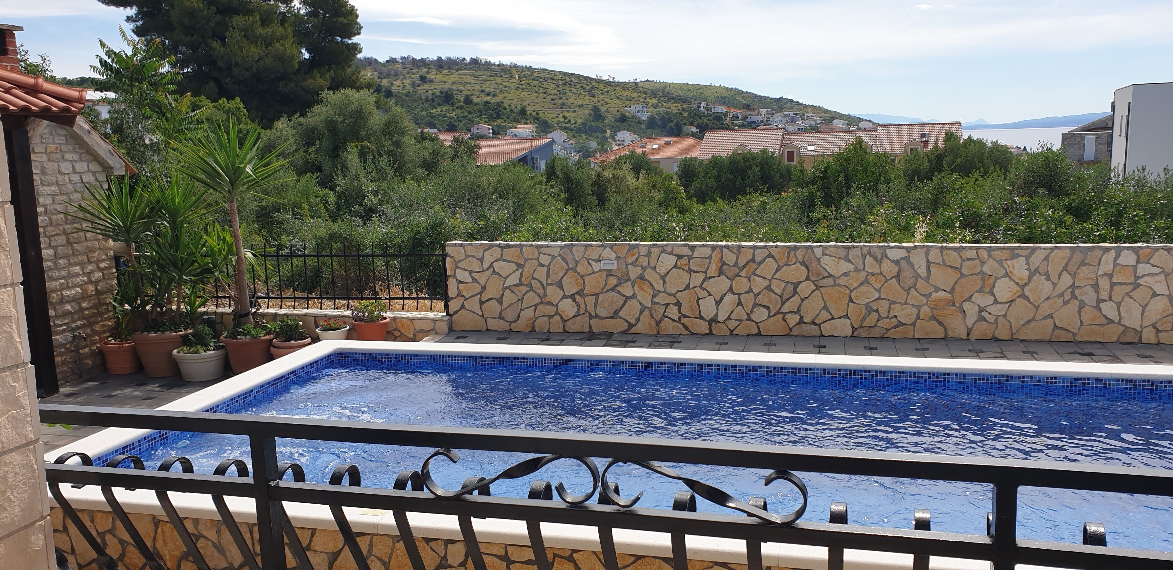 A swimming pool with a stone wall and metal railing, surrounded by potted plants and overlooking a hillside with houses.