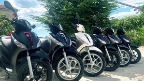 En række scootere parkeret udendørs med træer og en klar himmel i baggrunden.
