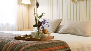 Una cama bien hecha con colcha, una mesita de noche con lámpara, una bandeja con frutas y un jarrón de flores.