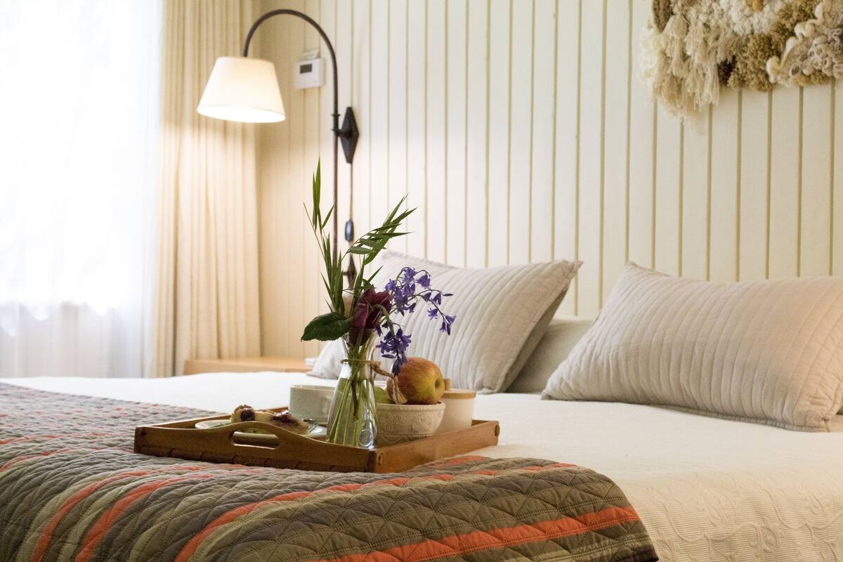 A neatly made bed with a quilt, a bedside table with a lamp, a tray with fruits and a vase of flowers.