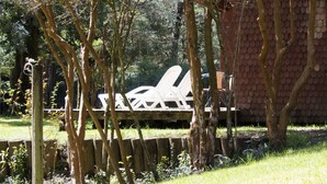 Una terraza de madera con sillas blancas, rodeada de árboles y un muro de ladrillo.