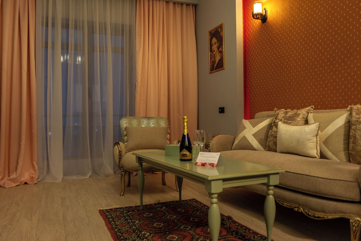 Una sala de estar con un sofá, una silla, una mesa con una botella y un vaso, un tapete y una pared con una lámpara y un cuadro enmarcado.