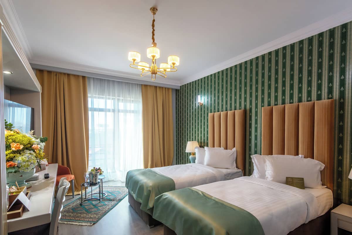Una habitación de hotel con dos camas, un escritorio con silla, una araña y un ventanal con cortinas.