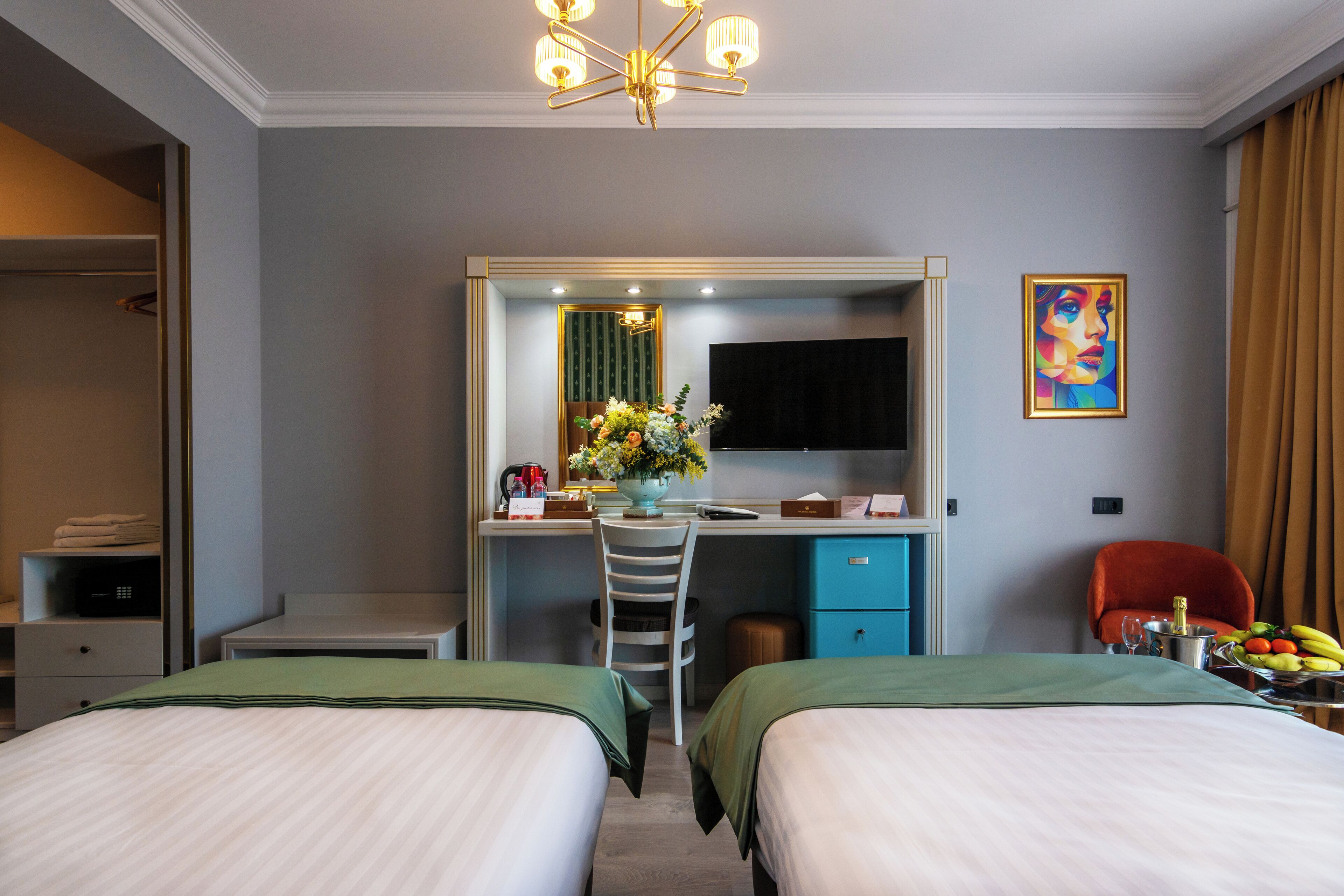 Habitación de hotel con dos camas, un escritorio con silla, un espejo, un televisor y un cuadro en la pared.