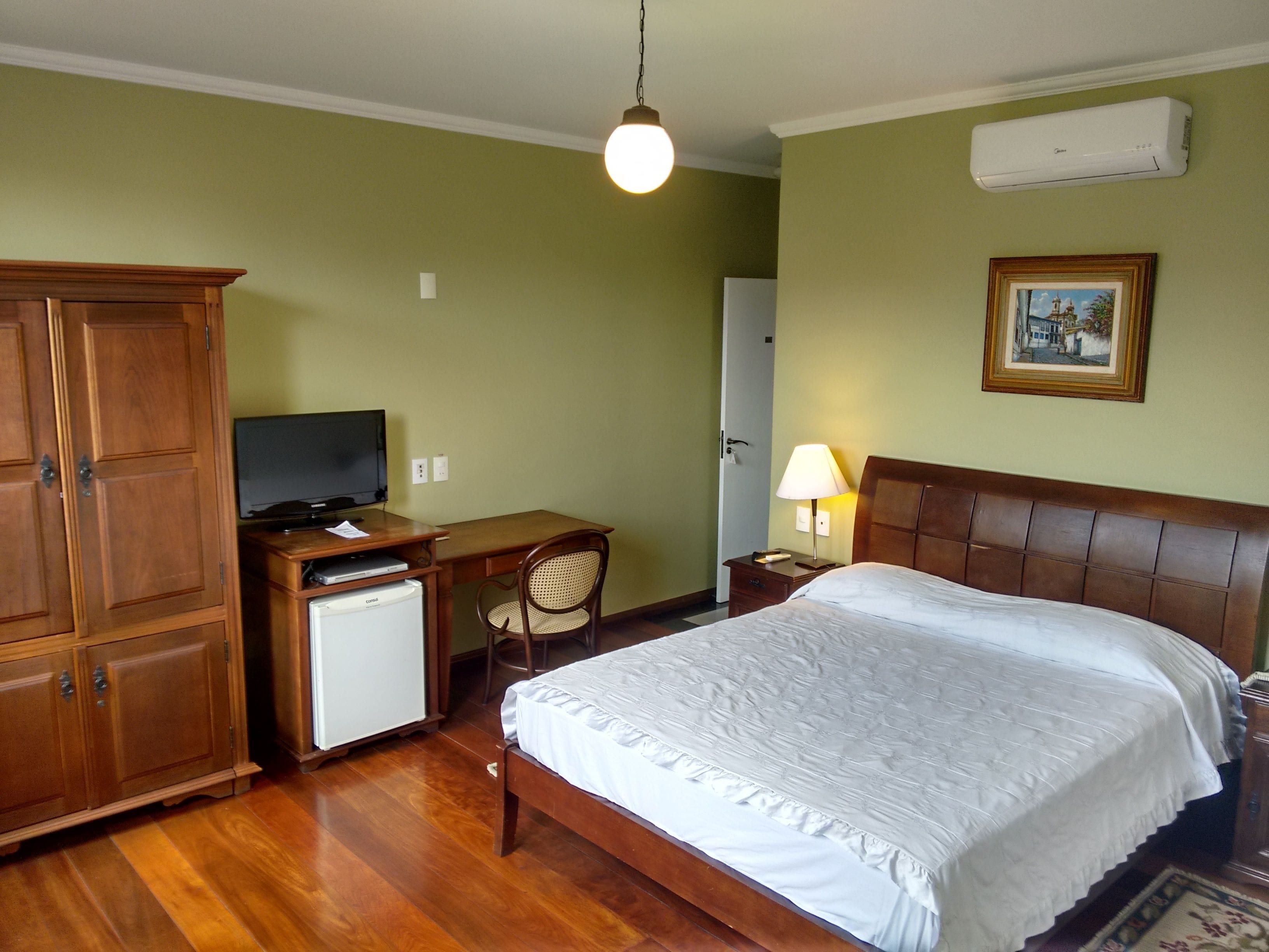 A hotel room with a wooden bed, a desk with a television, a small refrigerator, and a lamp.