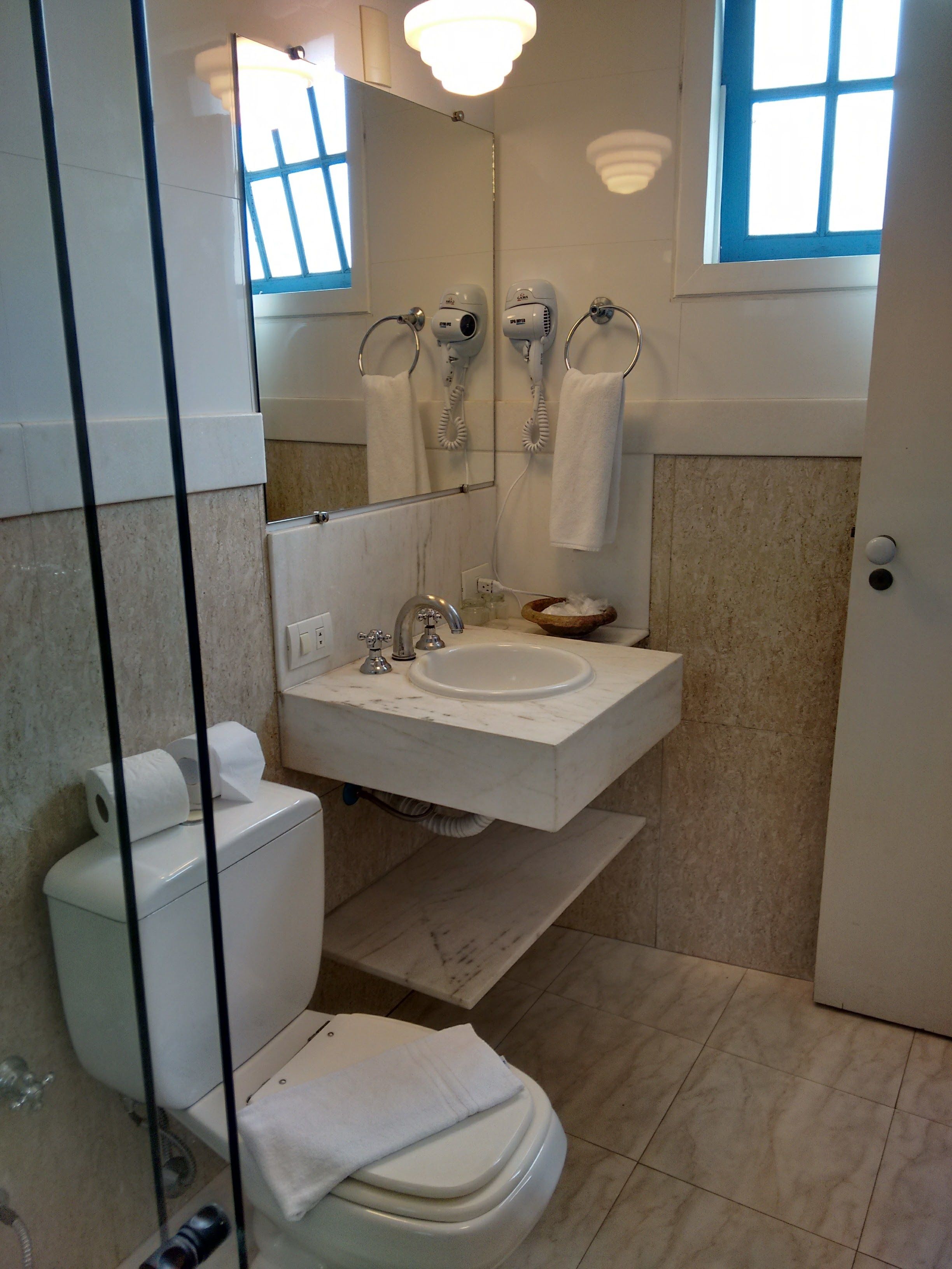 A bathroom with a toilet, sink, and mirror. There are two hair dryers on the wall, and a window above the mirror.