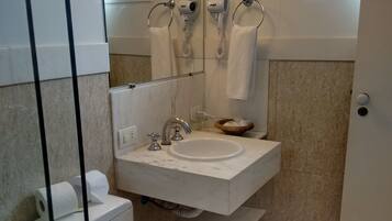 A bathroom with a toilet, sink, and mirror. There are two hair dryers on the wall, and a window above the mirror.