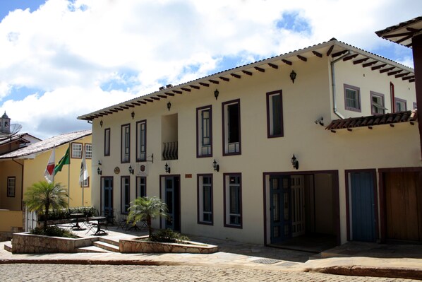 Un edificio con varias puertas y ventanas, un patio de piedra con plantas y una bandera.