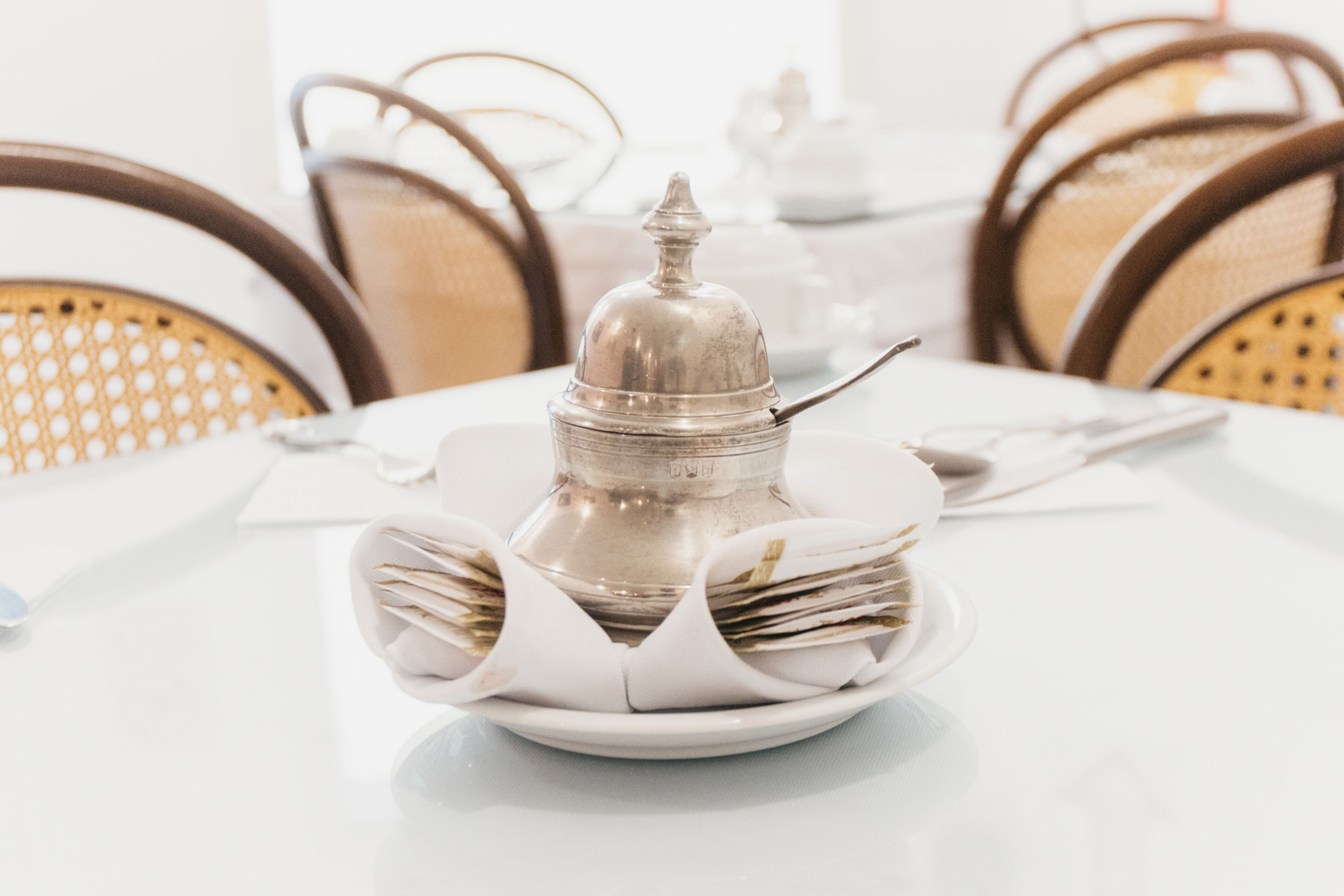 Uma mesa de jantar posta com uma chaleira de prata, uma pilha de jornais e um prato branco.