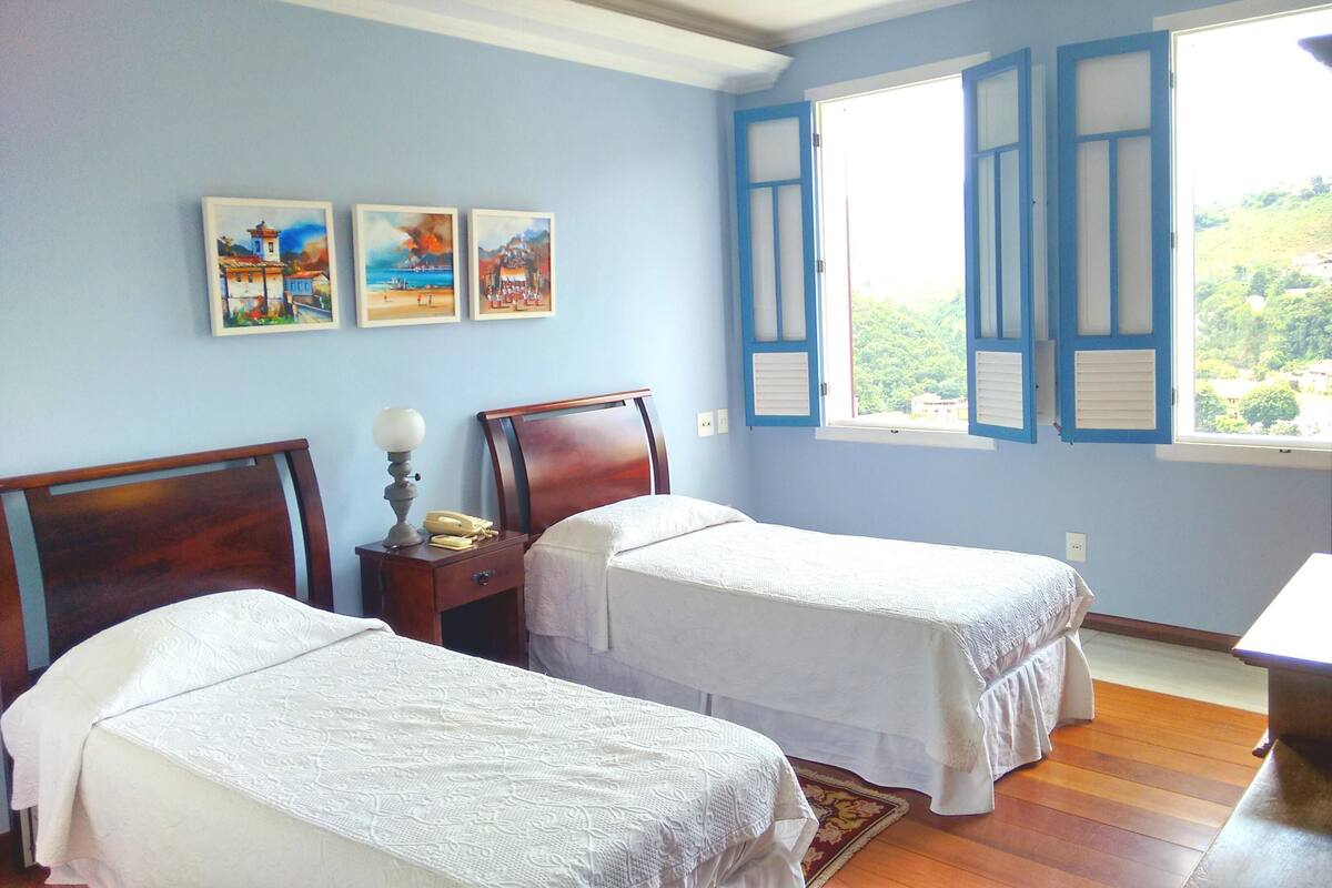 A room with two single beds, a nightstand with a lamp, and a window with shutters.