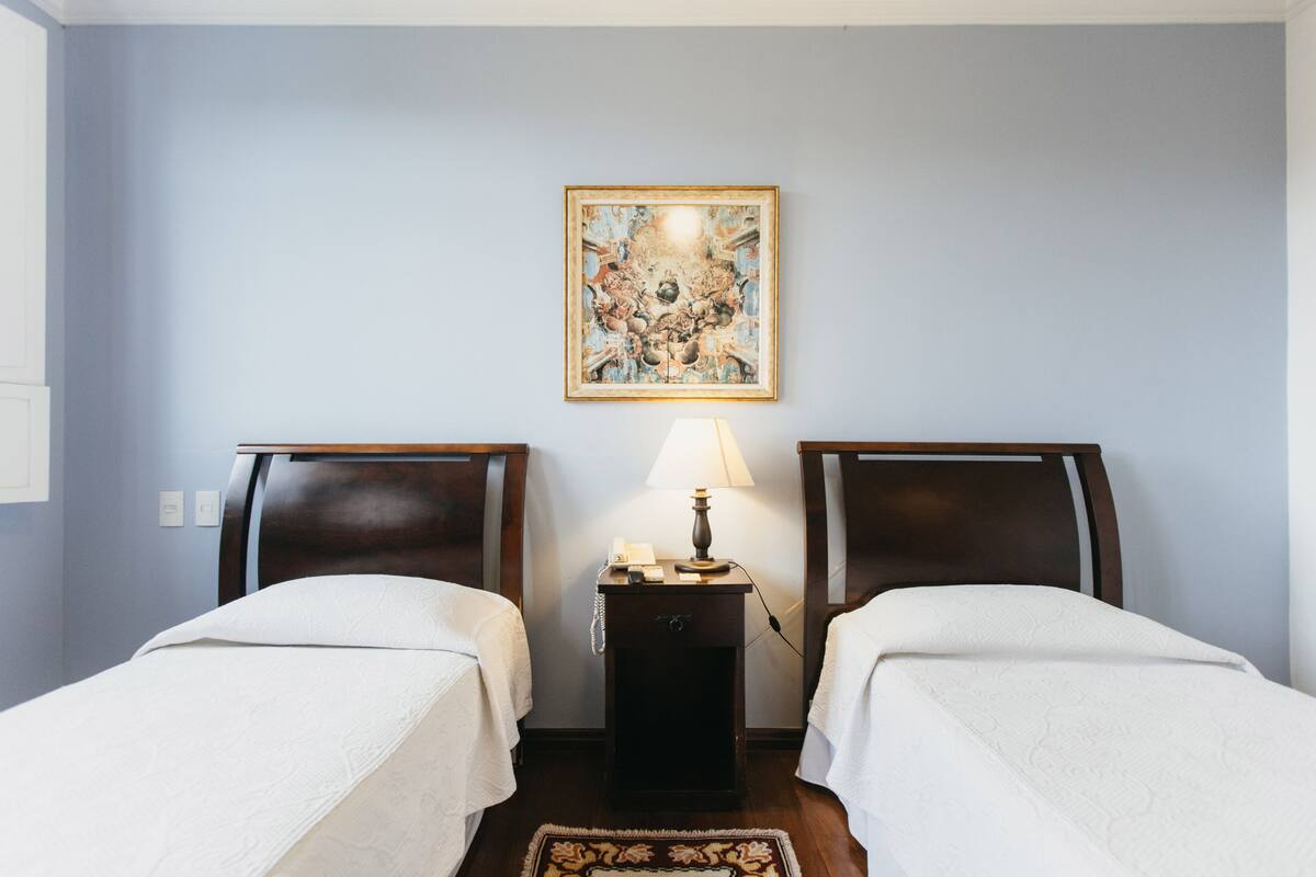 A hotel room with two single beds, a nightstand with a lamp, and a framed artwork on the wall.