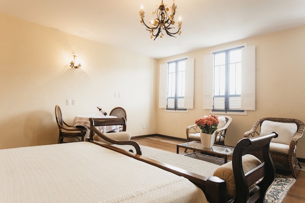 Habitación de hotel con una cama grande, dos sillones, una mesita, una araña y dos ventanas con contraventanas.