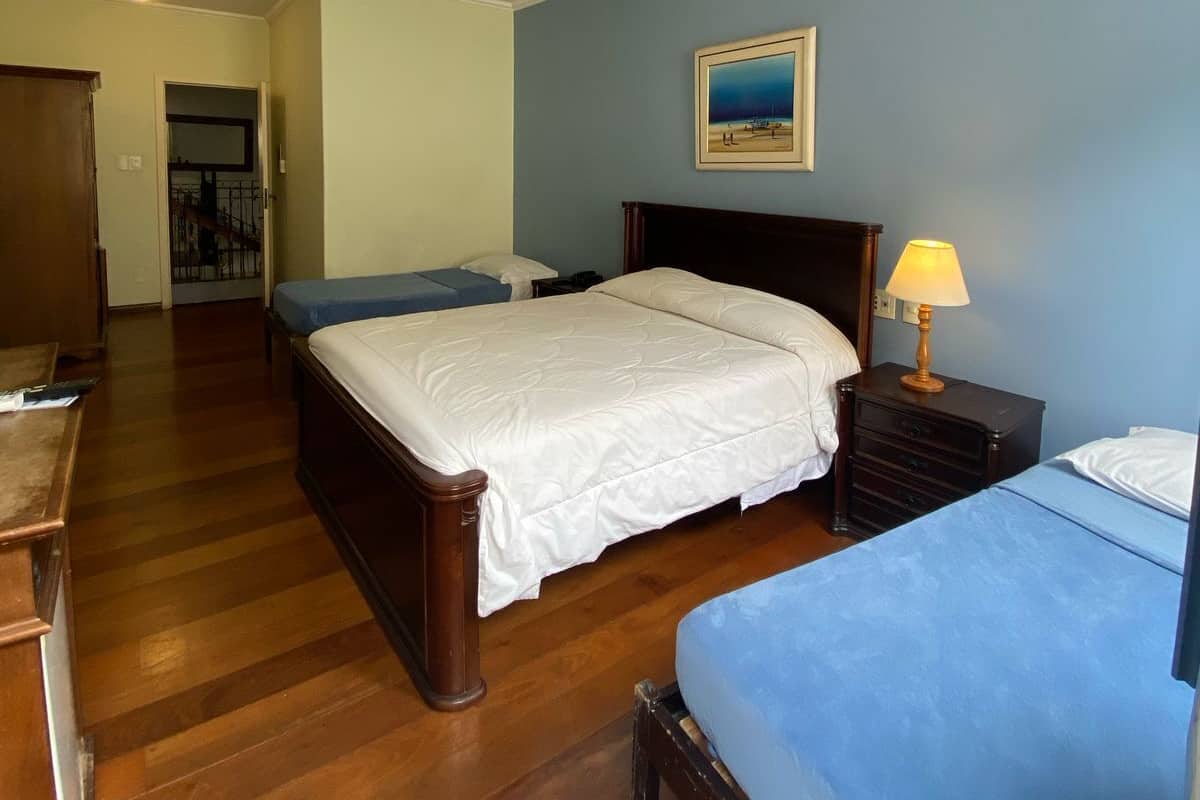 A bedroom with a large bed, a smaller bed, a desk, and a chair. There is a painting on the wall and a lamp on the desk.
