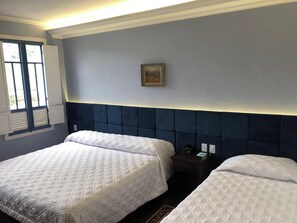 Habitación de hotel con dos camas, una mesita de noche, un cuadro enmarcado en la pared y una ventana con contraventanas.