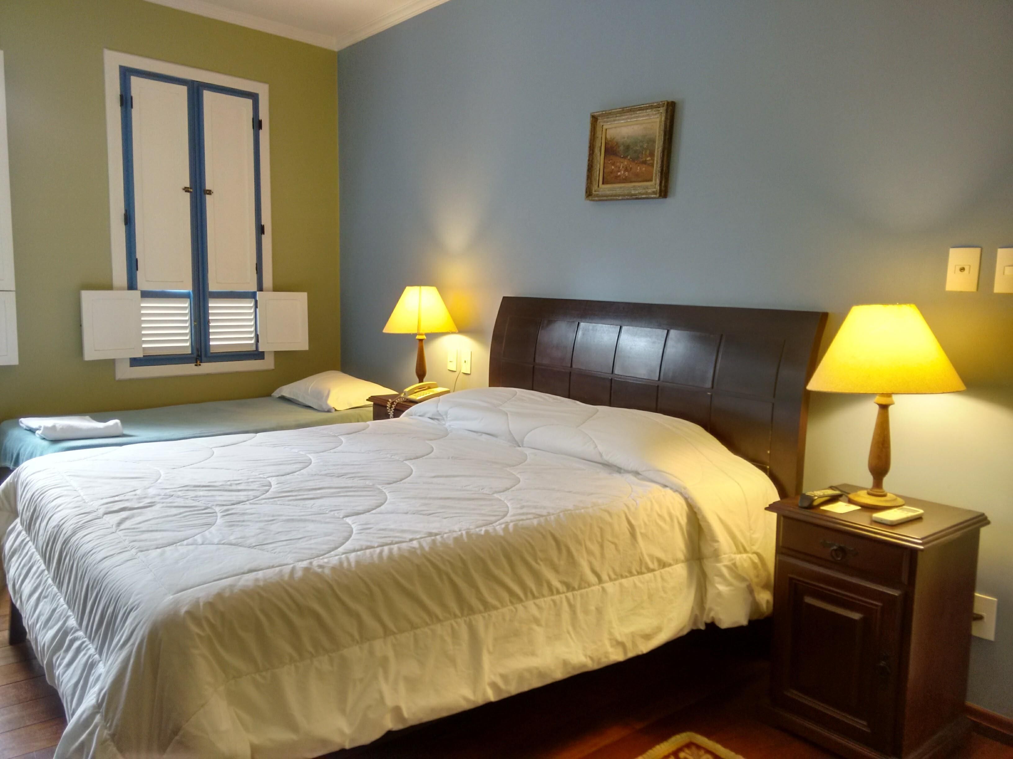 A hotel room with a large bed, two bedside tables with lamps, a nightstand, a wall-mounted telephone, and a window with shutters.