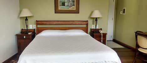 Una cama bien hecha con un cabecero de madera, dos mesitas de noche con lámparas, una silla y un cuadro en la pared.