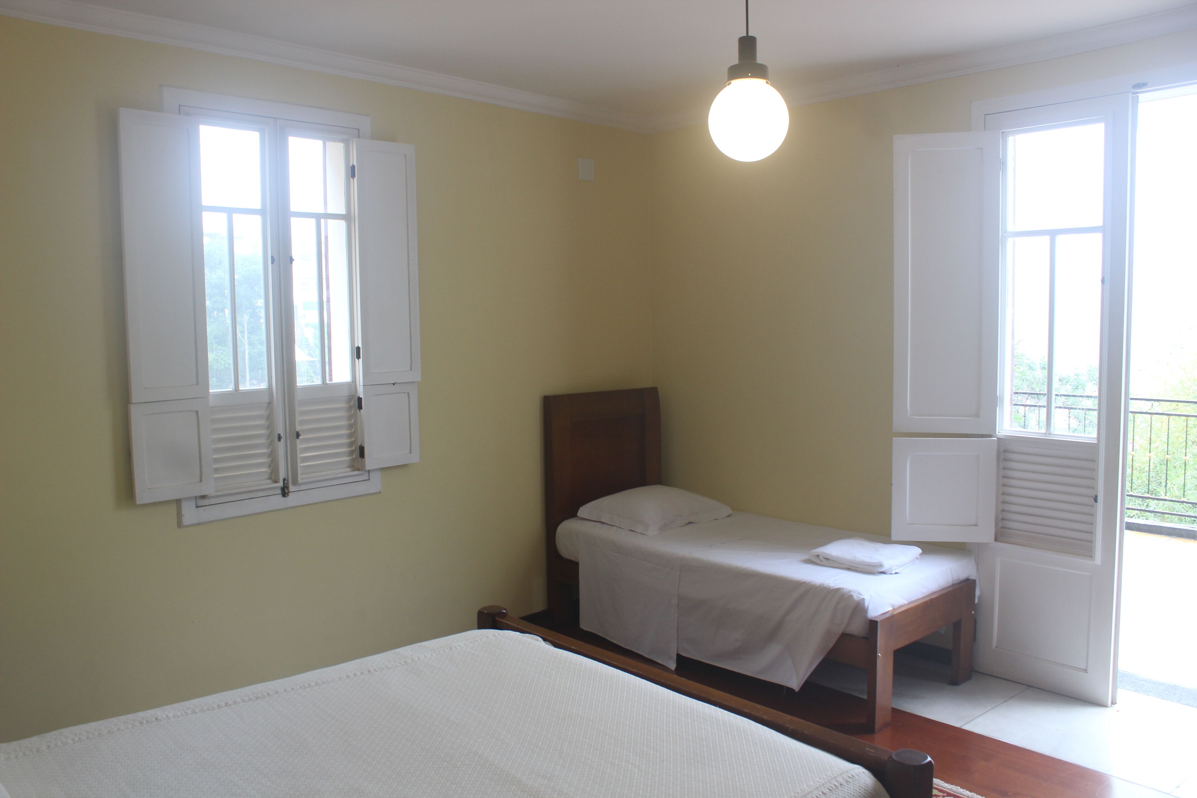 A bedroom with a single bed, a wooden headboard, a bedside table, and a window with shutters.