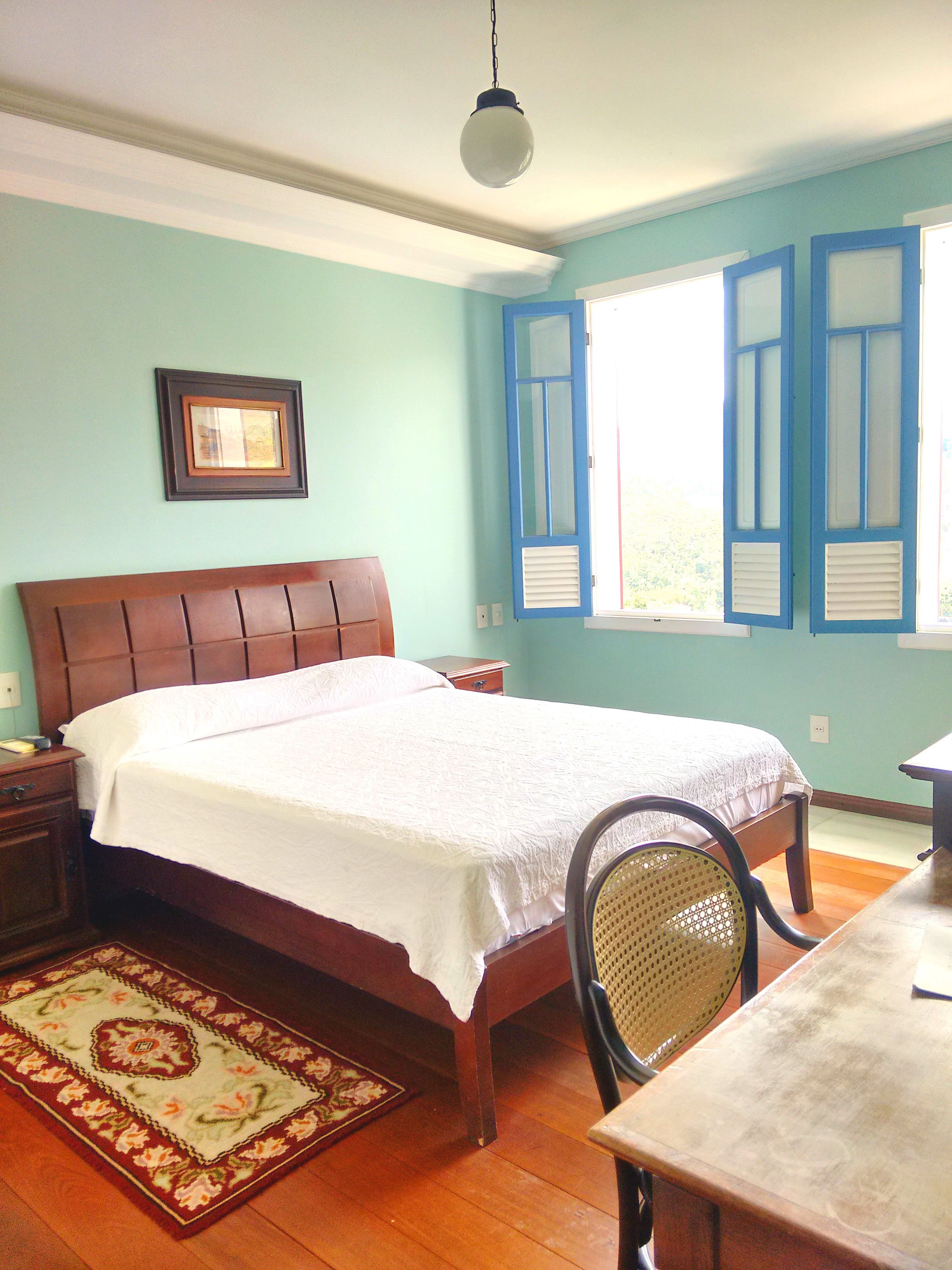 Un dormitorio con cama de madera, una mesita de noche, una silla, una mesa, un ventanal con contraventanas y una lámpara colgante.