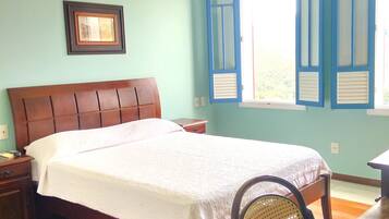 Un dormitorio con cama de madera, una mesita de noche, una silla, una mesa, un ventanal con contraventanas y una lámpara colgante.