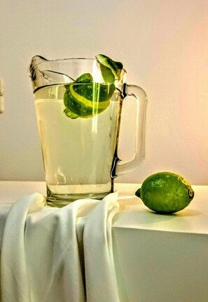 A clear glass pitcher with a handle, filled with a clear liquid, and a lime wedge inside. A whole lime is placed beside the pitcher. - Mykonos Chora Apartments (Mykonos)