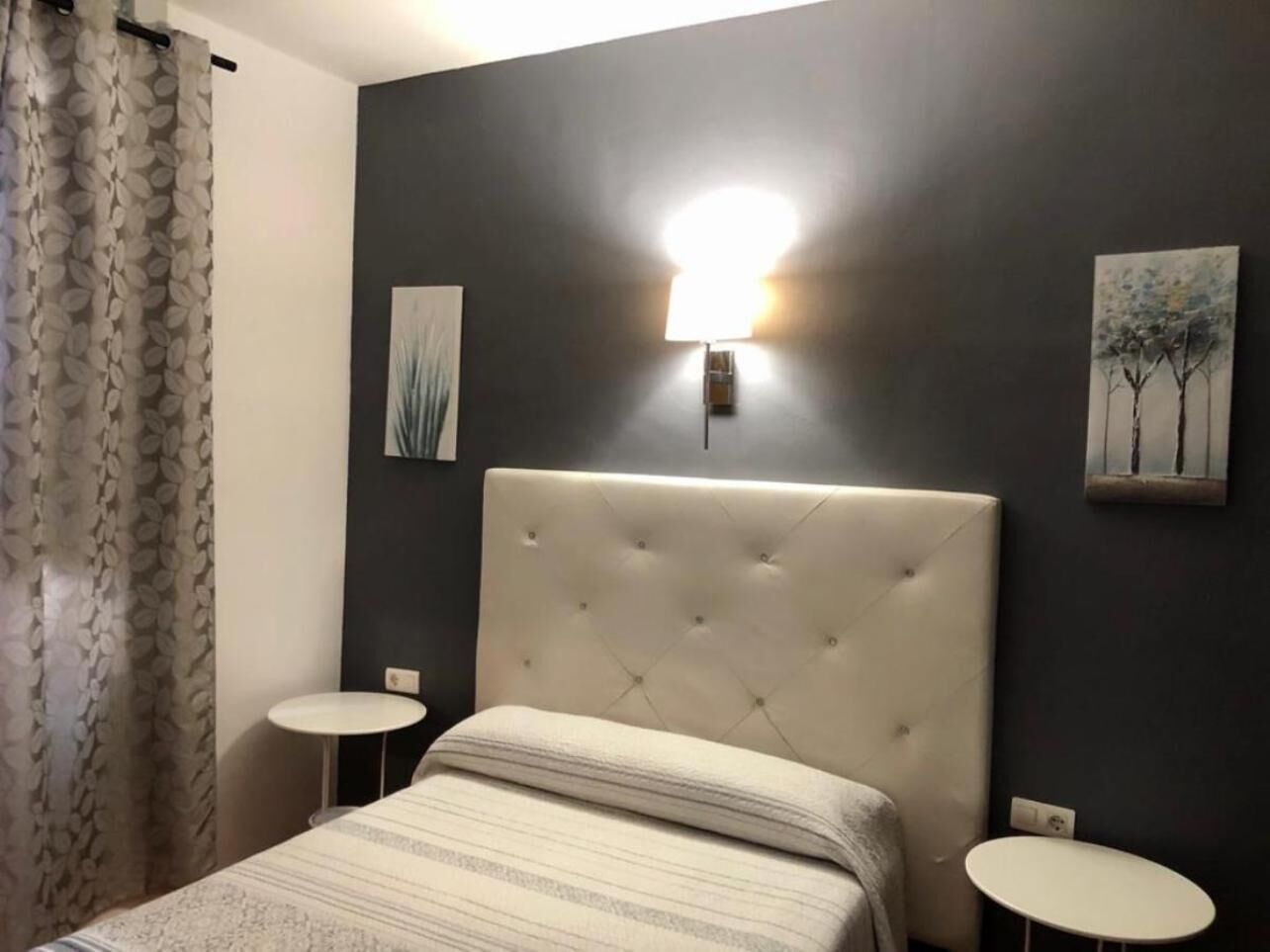 A bedroom with a tufted headboard, two bedside tables, a wall-mounted light, and two paintings.