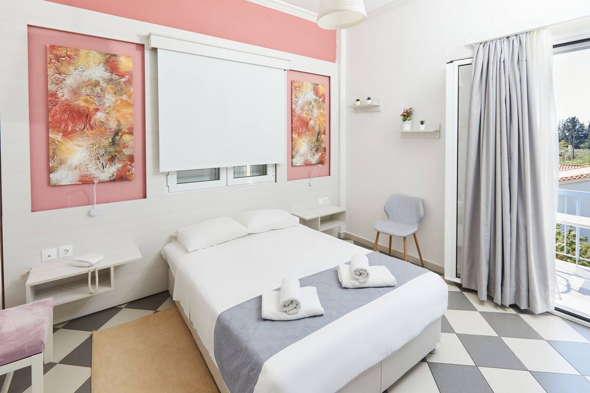 A modern hotel room with a large bed, two paintings on the wall, a small desk, a chair, and a balcony with curtains.