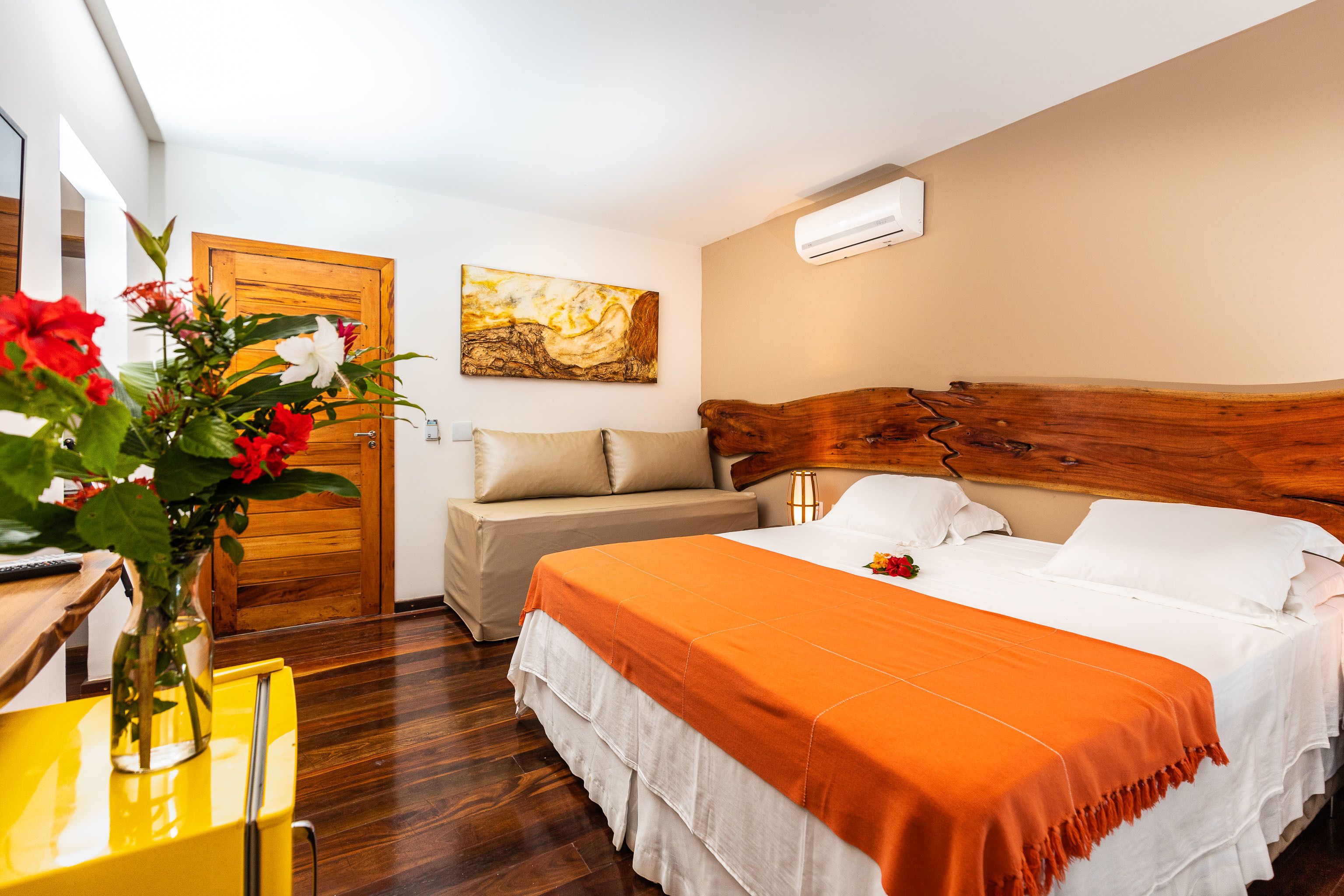 A hotel room with a wooden headboard, a bed with an orange bedspread, a beige sofa, a wooden door, and a painting on the wall.