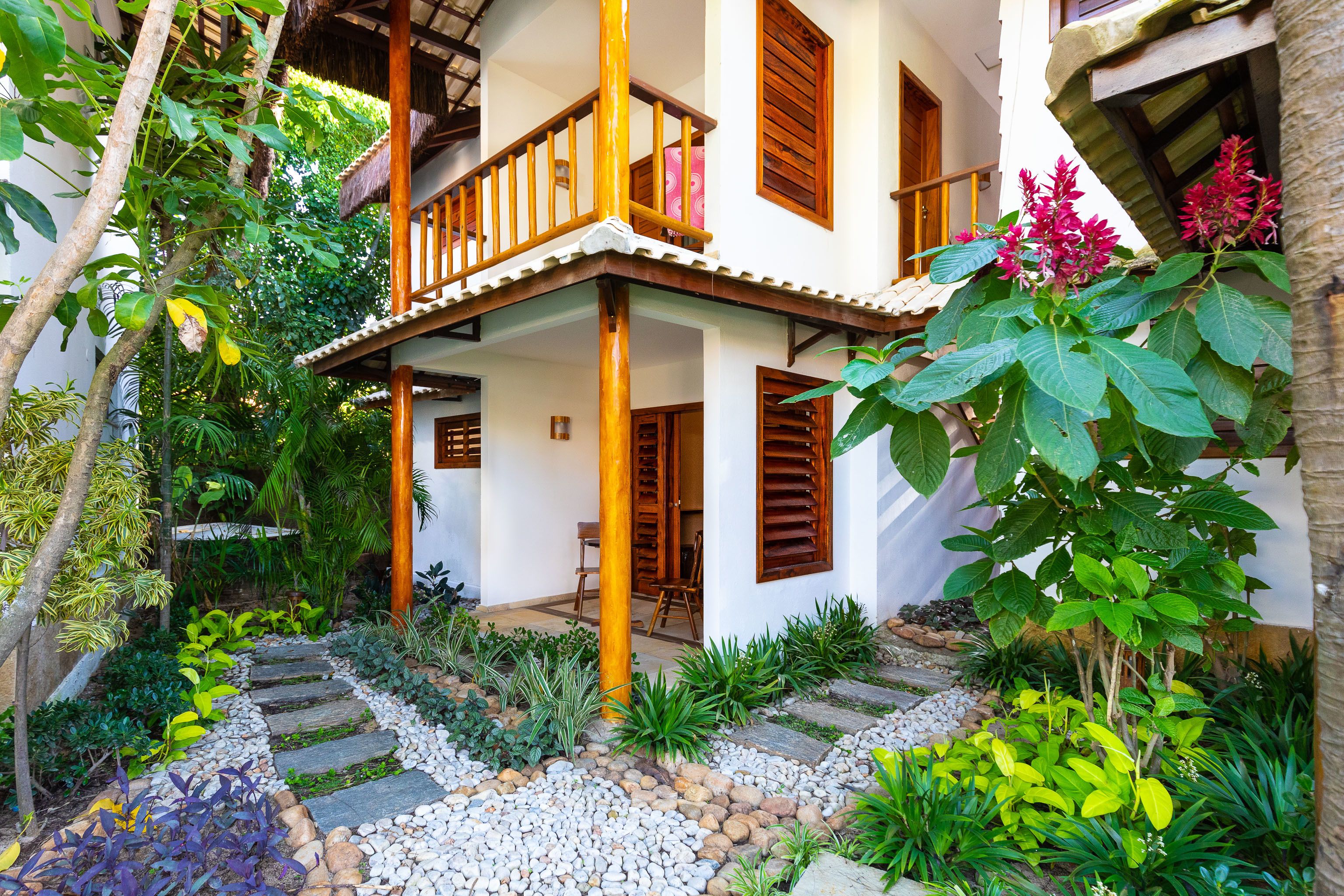 A two-story house with a balcony, surrounded by a garden with various plants and a stone pathway.