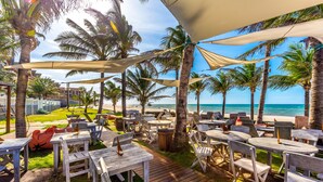 An outdoor seating area with wooden tables and chairs, surrounded by palm trees and overlooking the beach. - Vila Coqueiros (Caucaia)