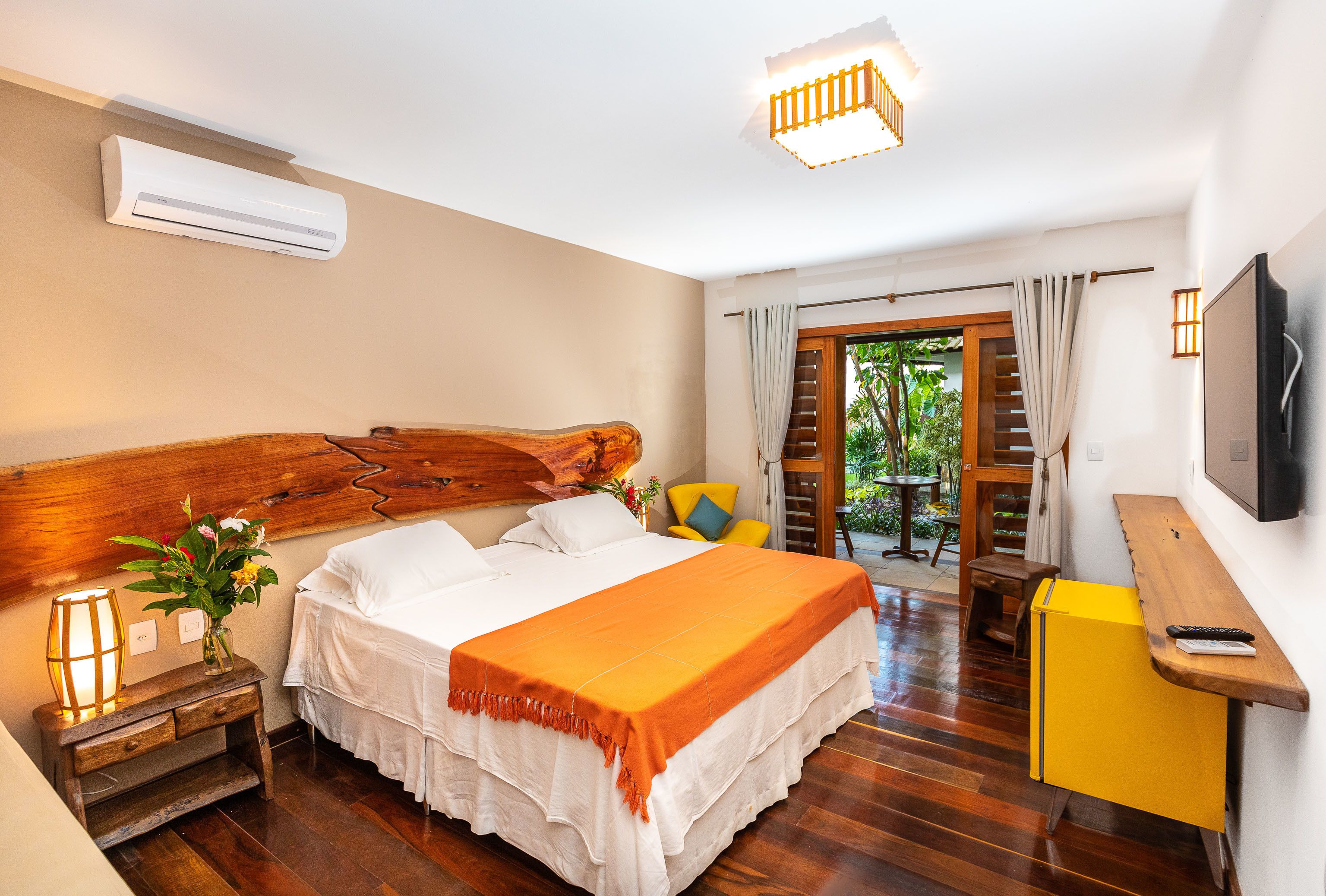 Une chambre à coucher avec une tête de lit en bois, un lit recouvert d’une couverture orange, une table de chevet avec une lampe, un petit bureau en bois et un téléviseur fixé au mur.