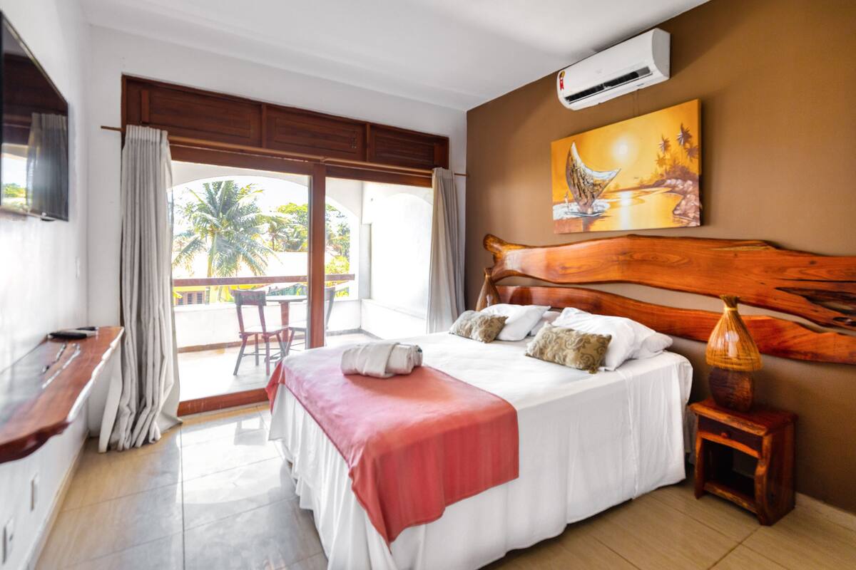 A hotel room with a large bed, a wooden headboard, a painting on the wall, and a view of palm trees through the balcony doors.