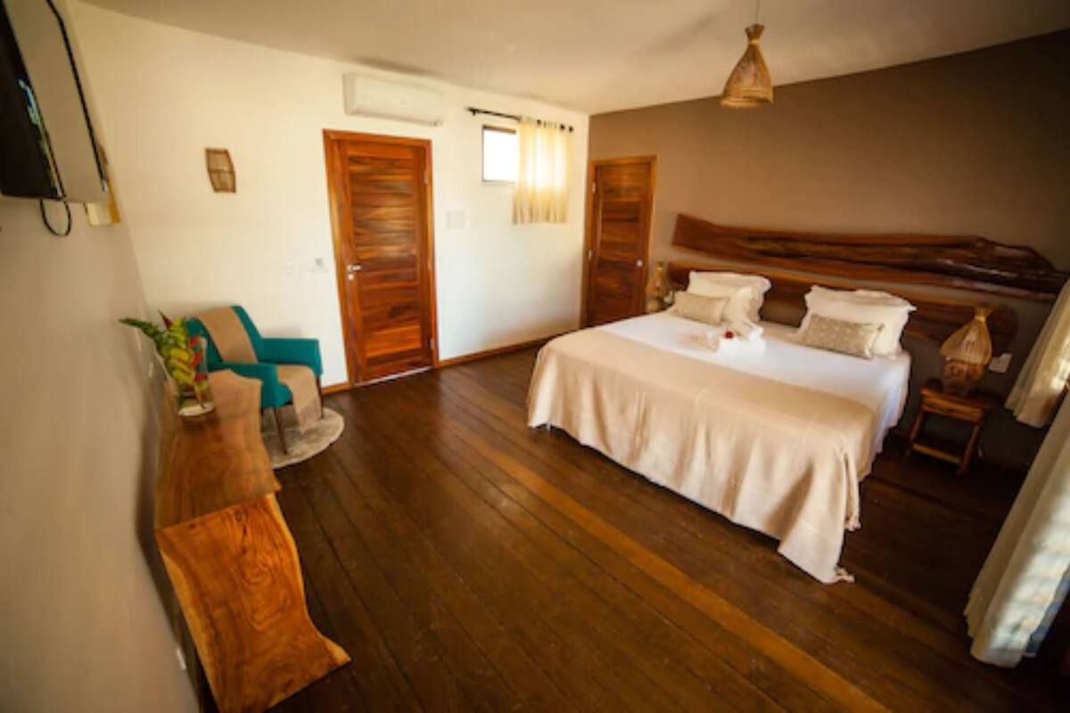 A bedroom with a large bed, wooden flooring, a wooden headboard, a small seating area with a chair and table, and a TV mounted on the wall.