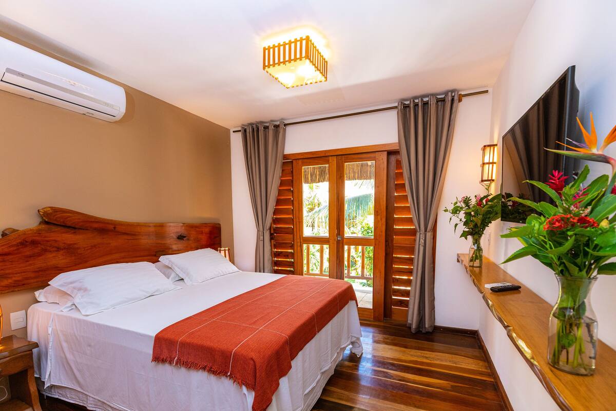 A bedroom with a wooden headboard, a large bed with white linens, a red throw blanket, a wooden floor, a large window with curtains, and a vase with flowers on a nightstand.